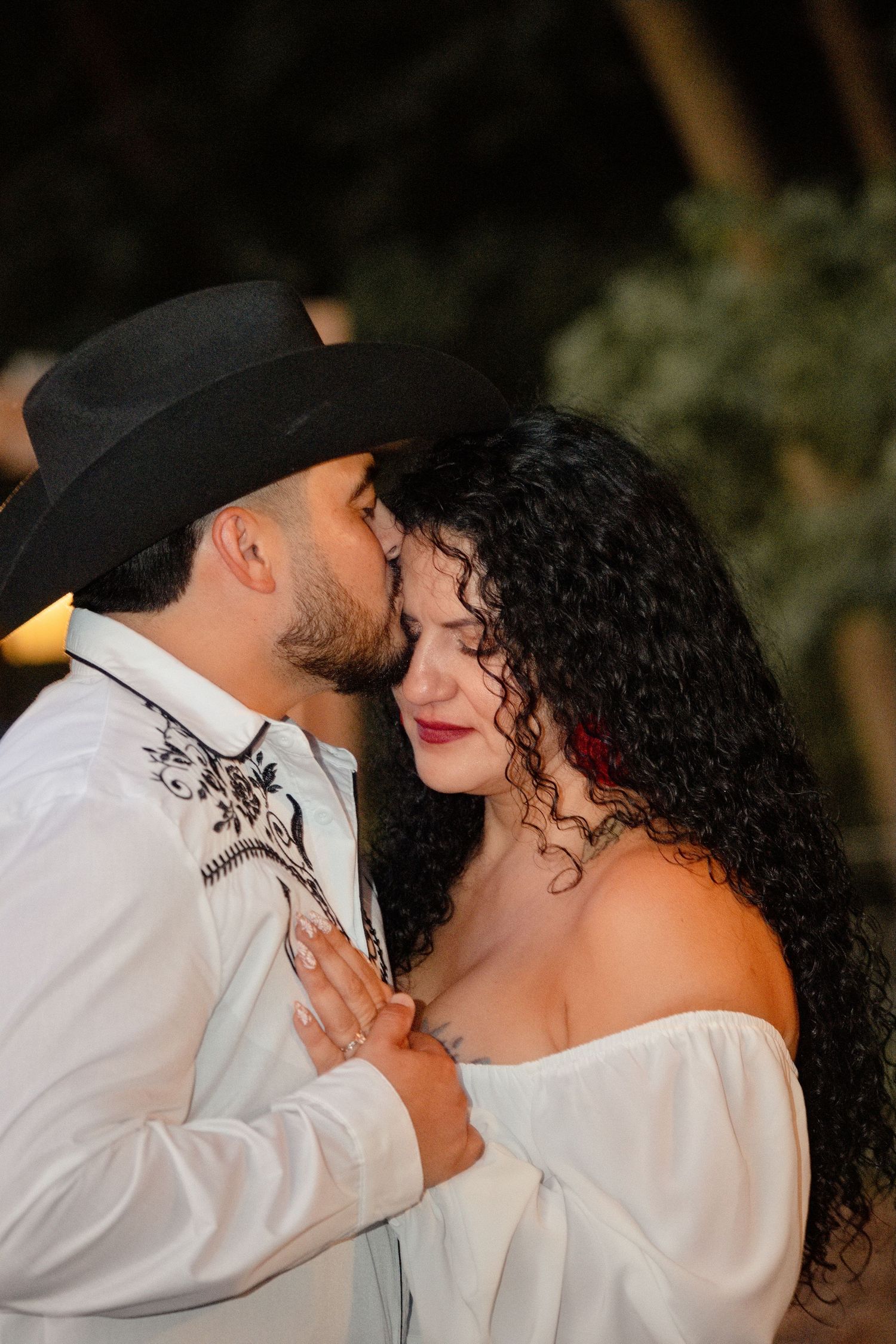 A man in a black cowboy hat and white embroidered shirt kisses a curly-haired woman in an off-shoulder white top.