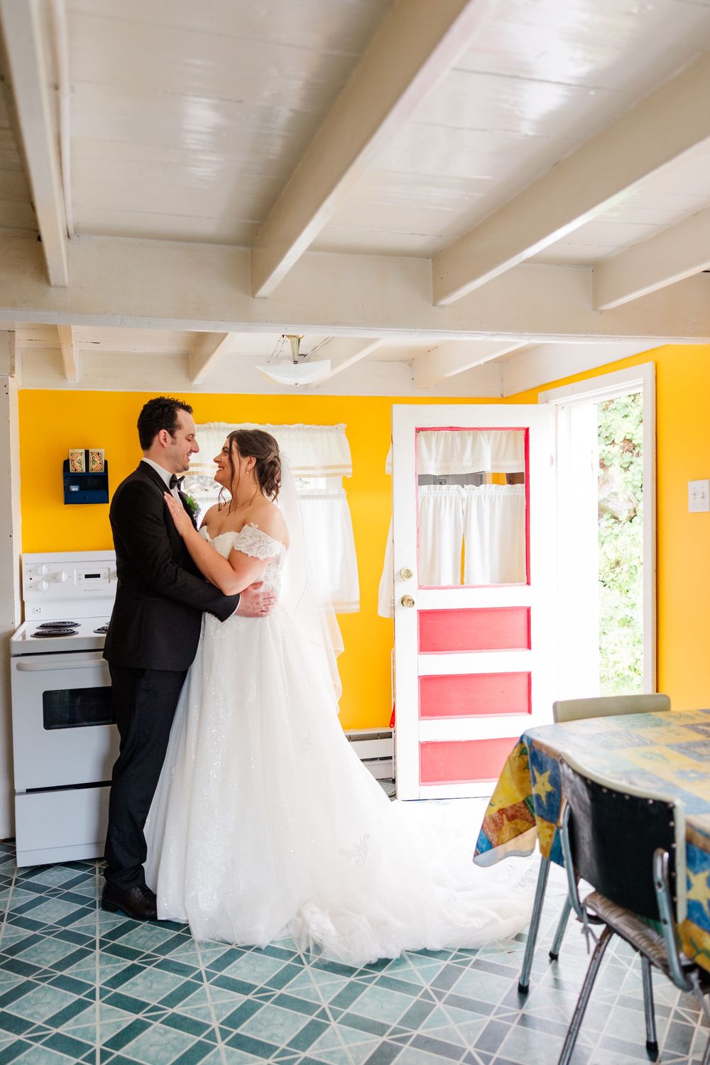 Bride and groom embracing in a vibrant yellow kitchen with teal tile floors and a colorful pink door on their wedding day.