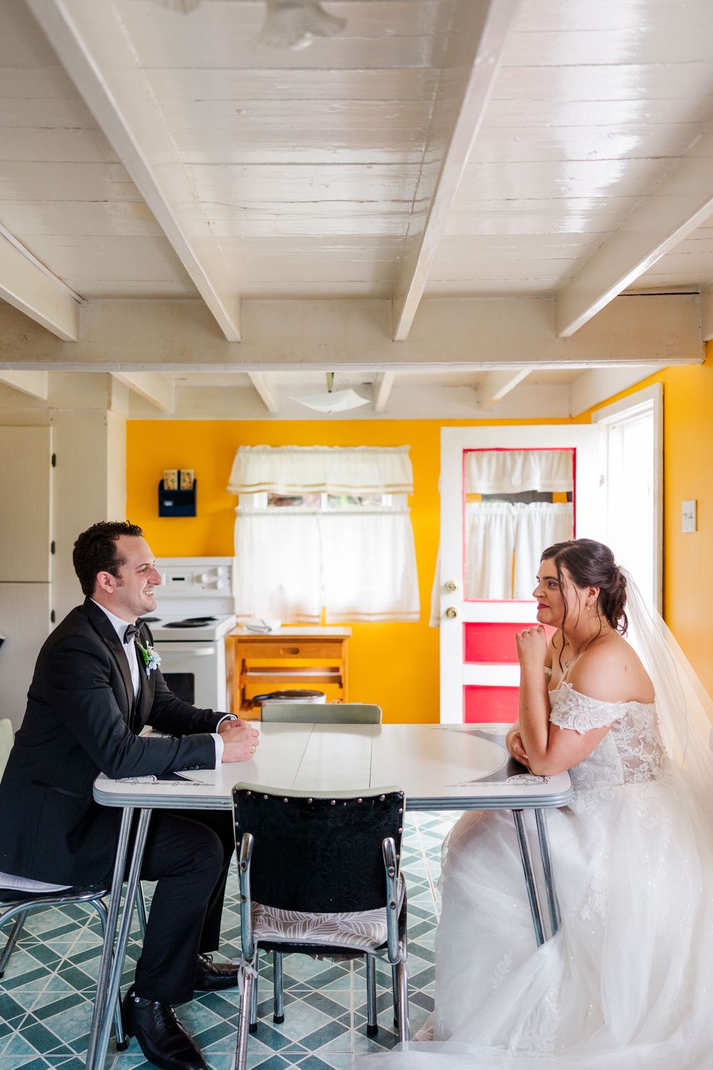 Bride and groom in wedding attire sit across from each other at a retro kitchen table with bright yellow walls.