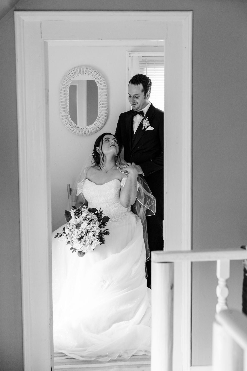 Black and white wedding photo of bride holding bouquet seated as groom in tuxedo stands behind her smiling indoors.