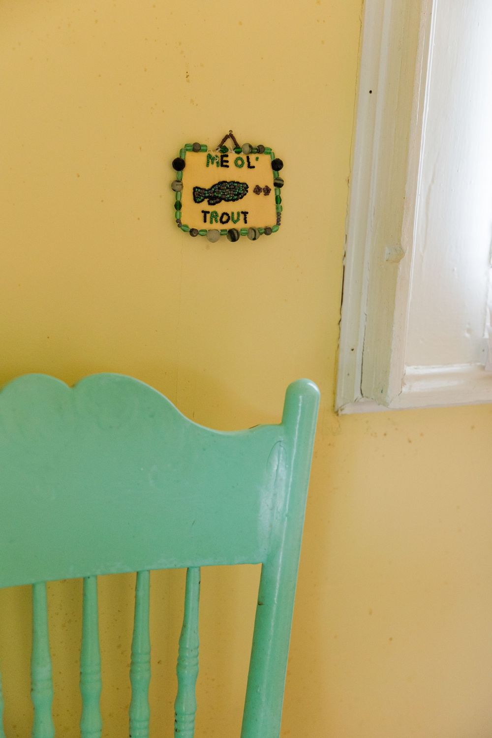 Turquoise wooden chair against a yellow wall with a decorative fish plaque reading Me Ol Trout near a white window.