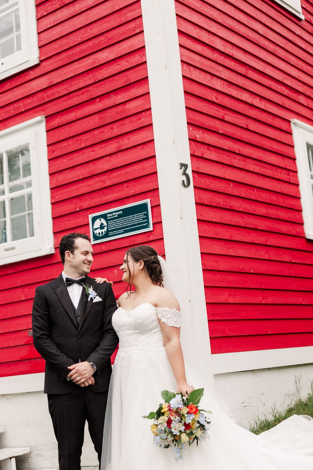 Bride and groom share a romantic moment beside a red building, bride holding a colorful bouquet.
