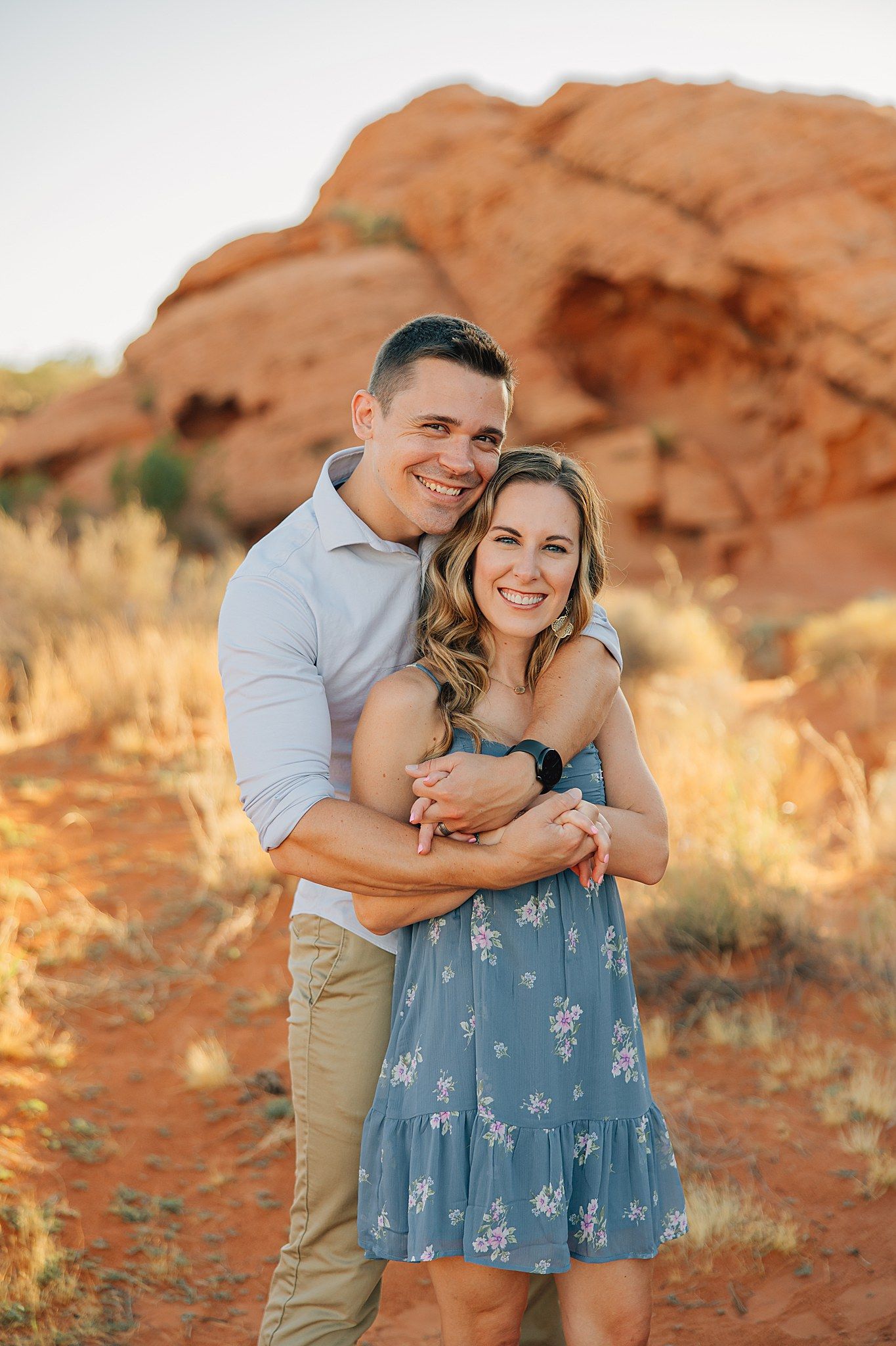 Couple embracing and smiling in red rock desert landscape, woman in blue floral dress, man in white shirt.