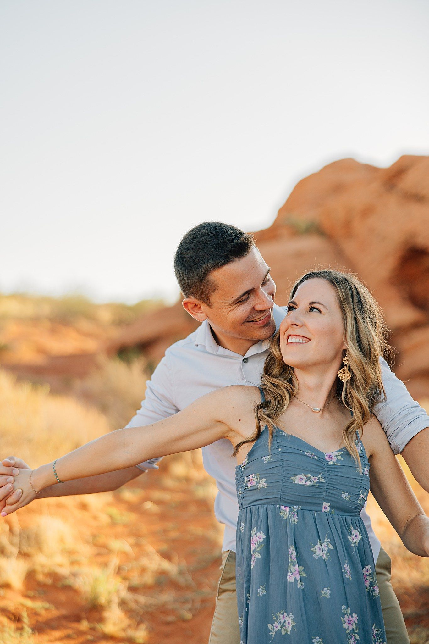 Smiling couple embracing outdoors in red rock desert landscape during golden hour engagement photo session.