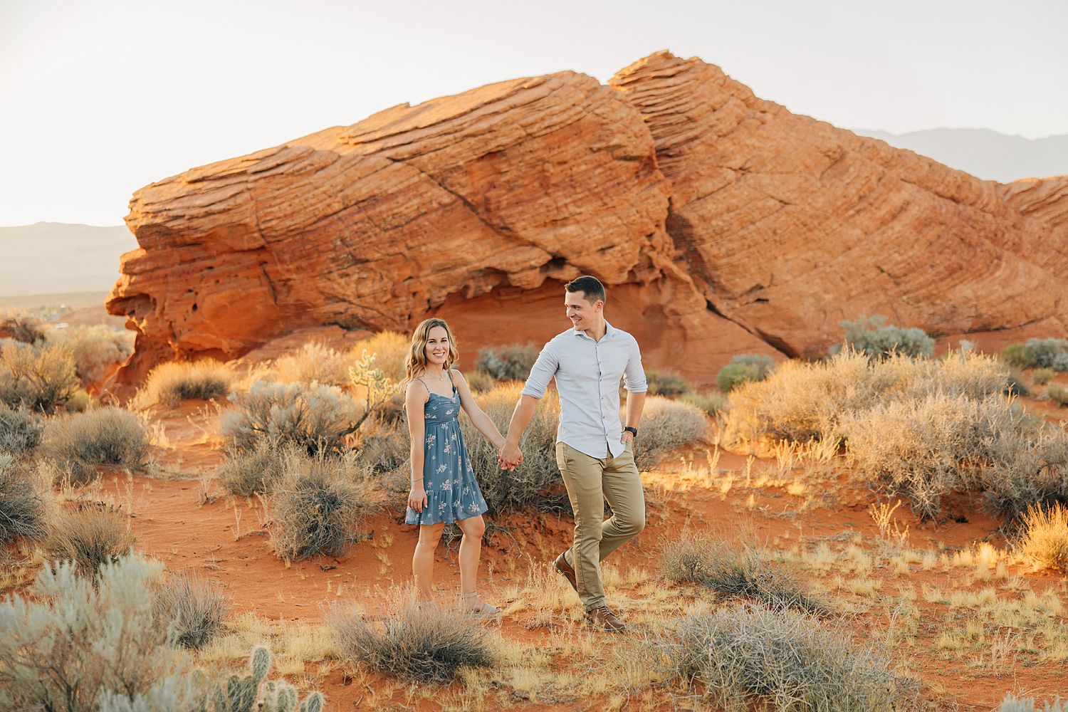 Couple holding hands walking through red rock desert landscape with sandstone formations and desert shrubs at golden hour.