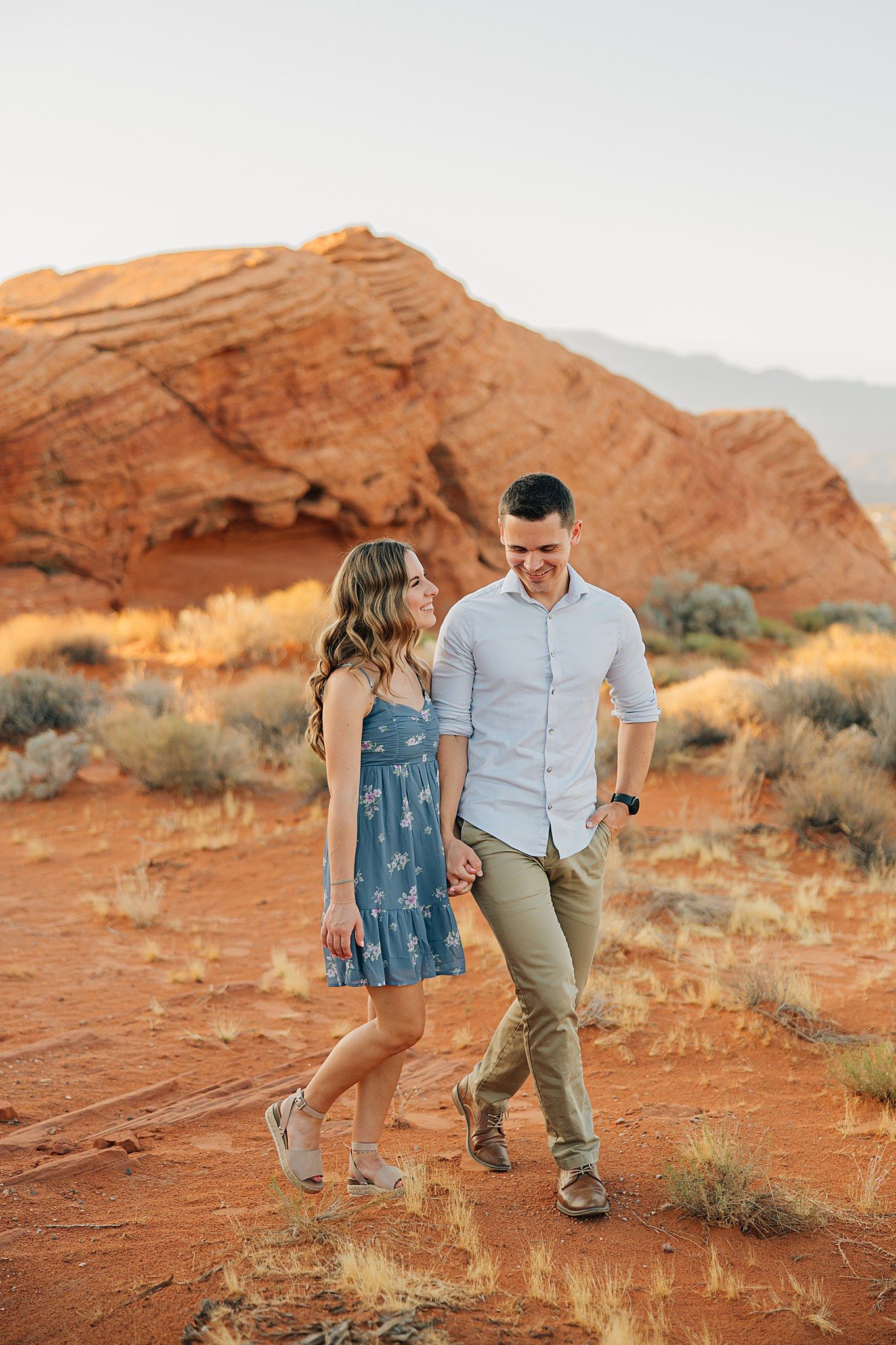 Couple holding hands walking on red desert sand with large sandstone rock formations at golden hour.