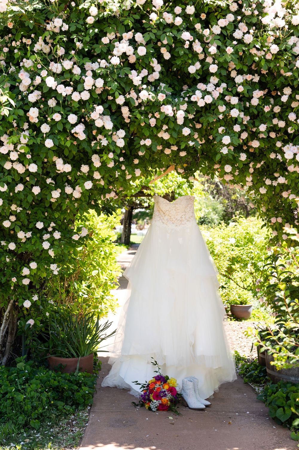 Wedding dress hanging on a floral arch with white roses, white cowboy boots and colorful bouquet below.
