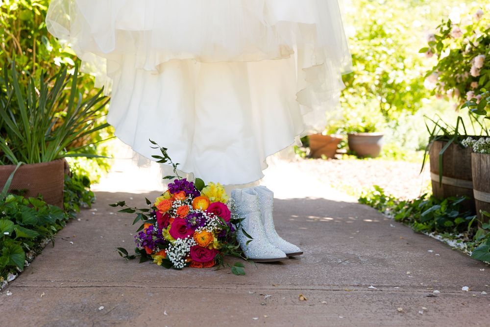 Bride in white gown standing on a garden path with colorful bouquet and white boots at her feet.