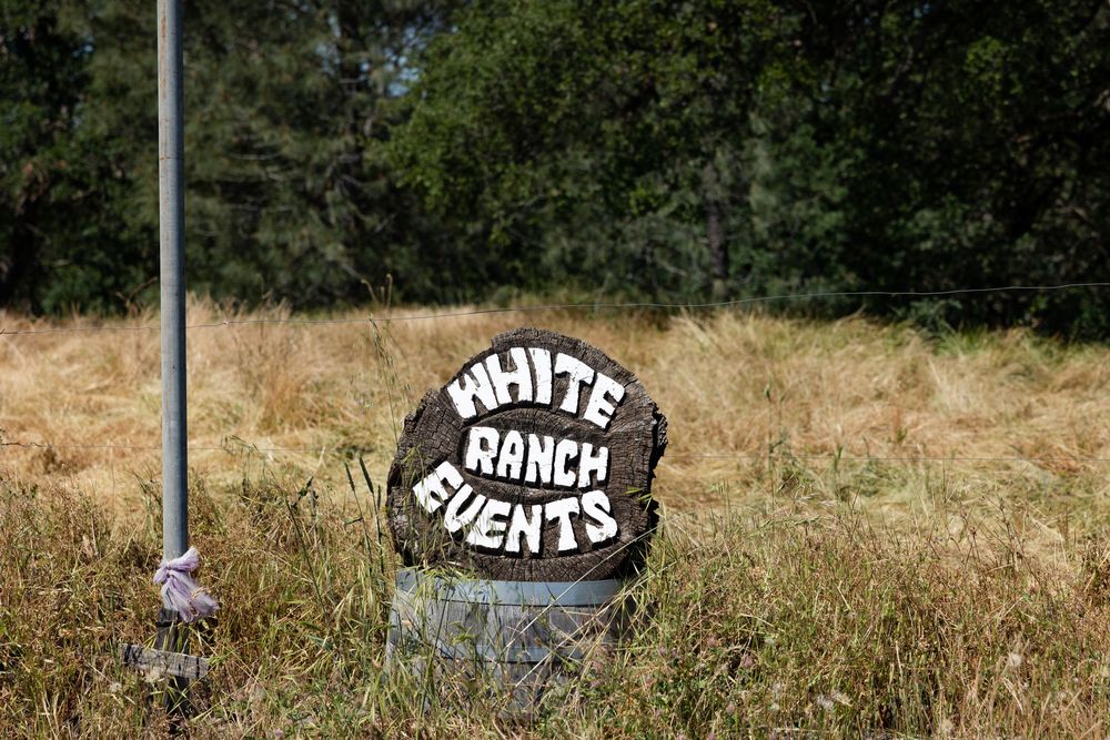 Rustic log sign reading White Ranch Events surrounded by dry grass and trees.
