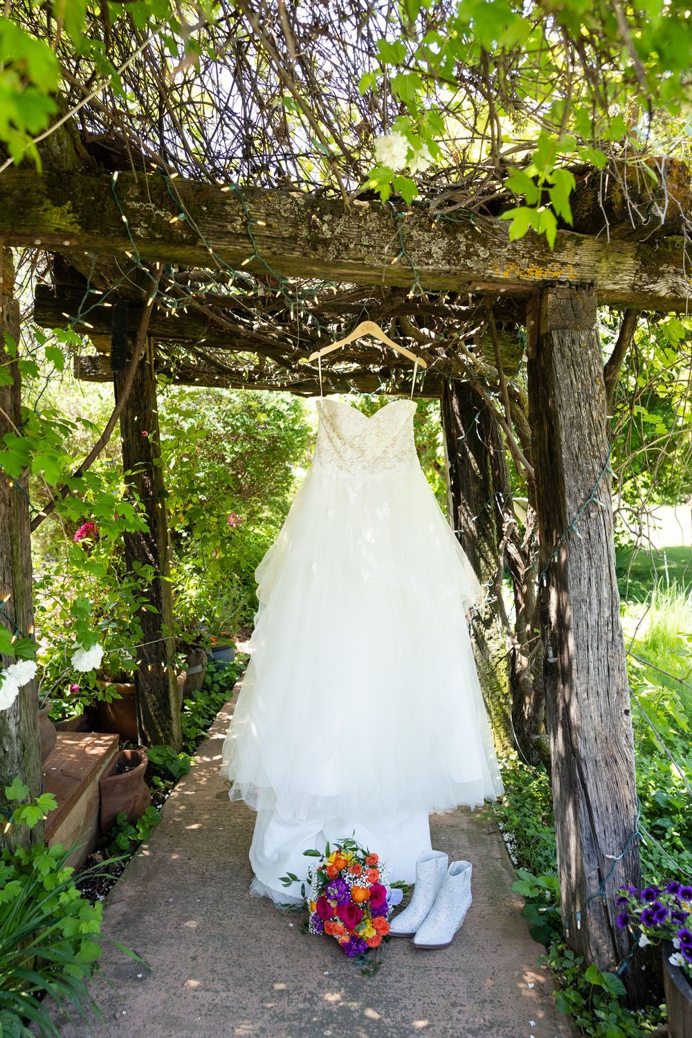 A white strapless wedding gown hangs on a rustic wooden arbor with a colorful bouquet and white shoes below.