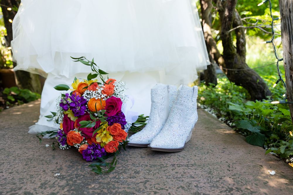 Colorful bridal bouquet with orange and purple flowers beside white glitter boots and wedding dress.
