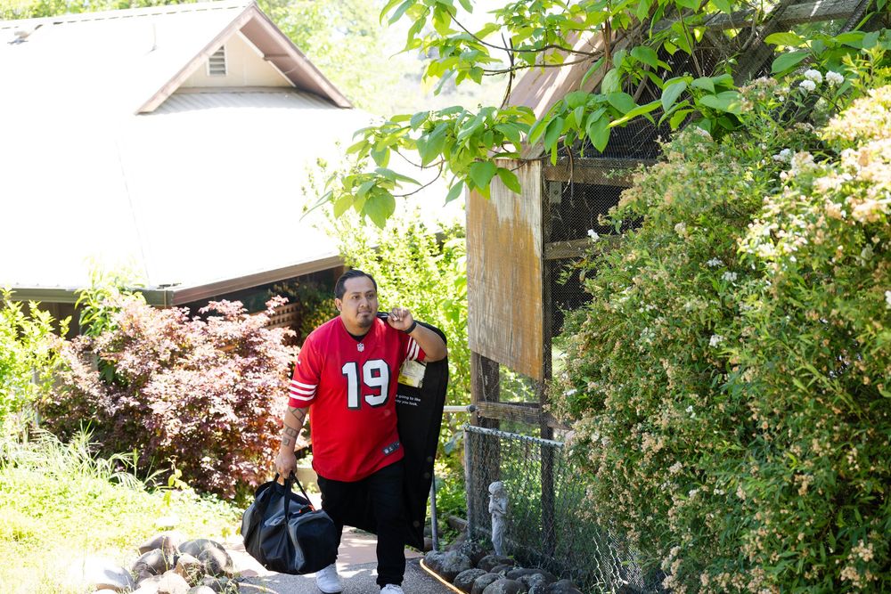 Person wearing a red number 19 jersey carrying a bag outside a house surrounded by lush green trees.