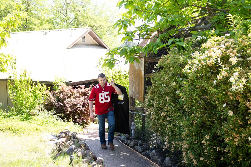 Man in red jersey number 85 walking along a garden path beside a rustic building.