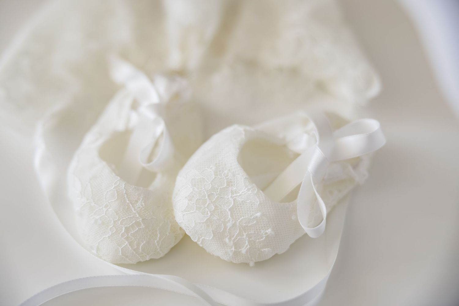 Delicate white lace baby shoes with satin ribbon ties resting on soft white fabric.