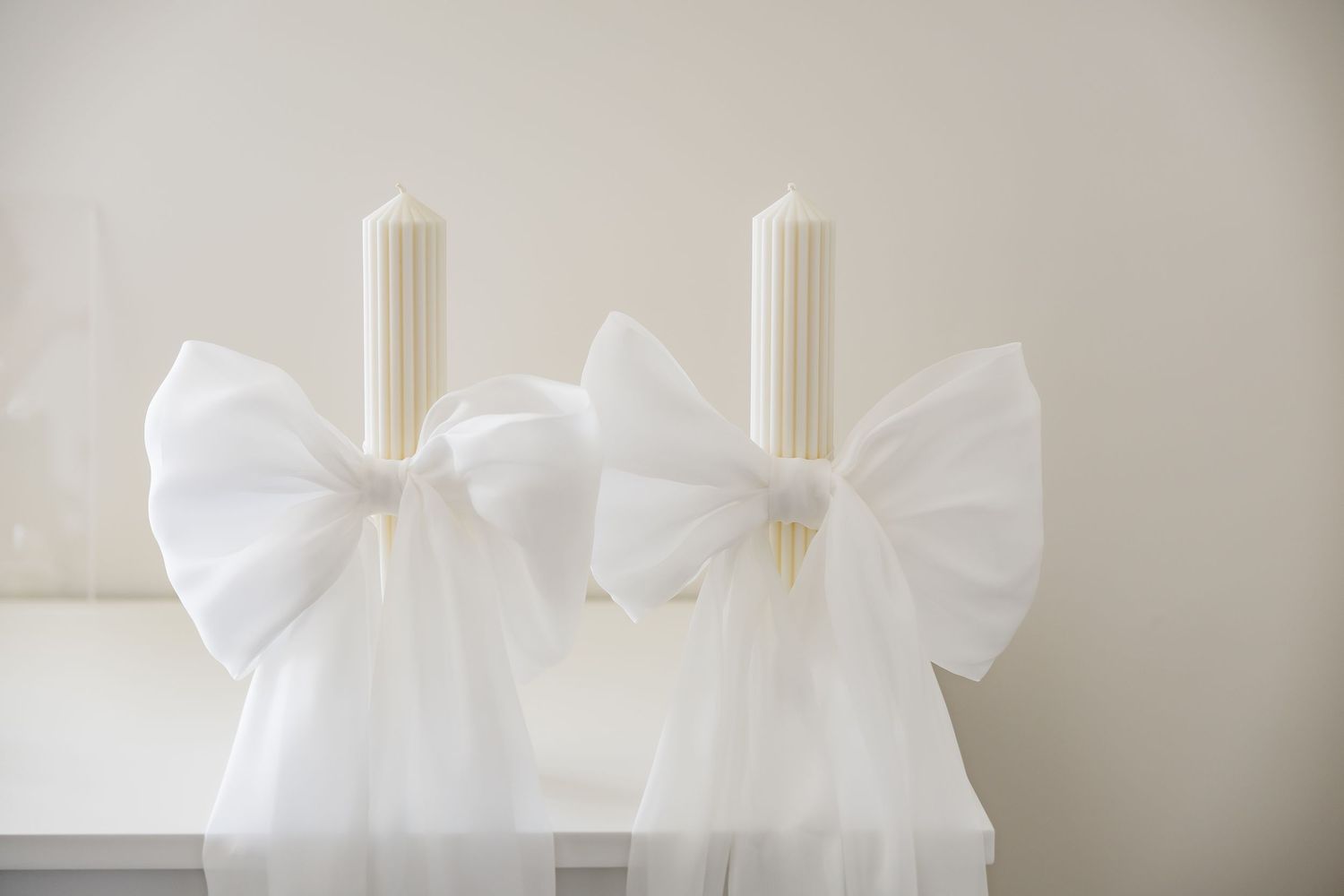 Two white pillar candles adorned with large white tulle bows displayed on a white surface.