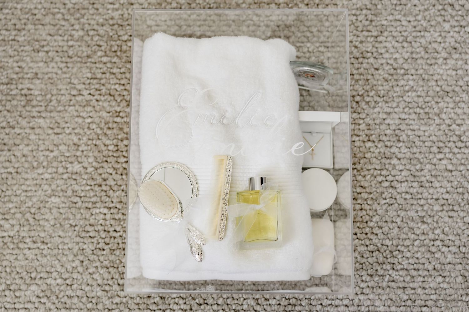 Christening flat lay with white gown, perfume bottle, cross necklace and accessories on neutral textured surface.