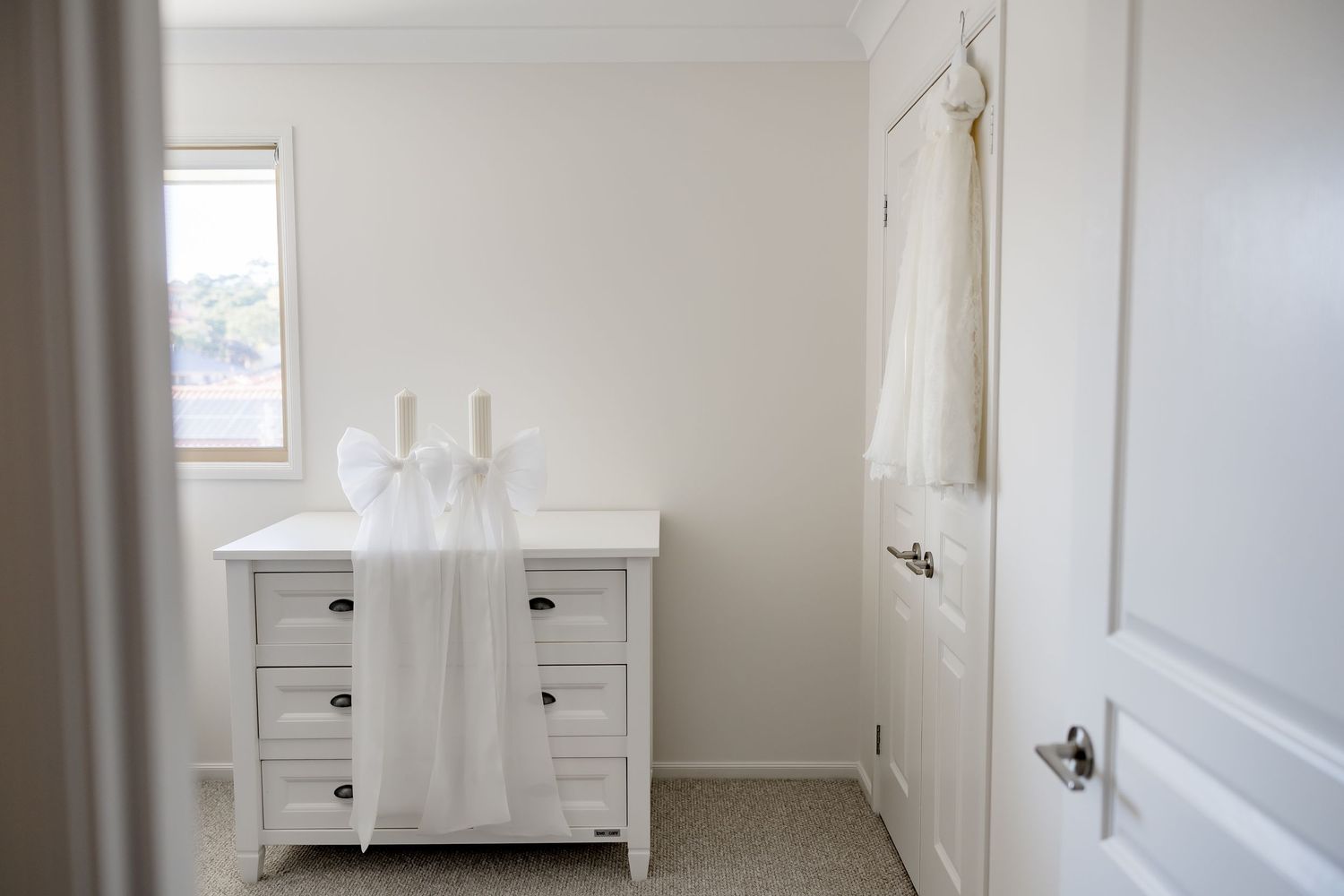 Bright white nursery room with dresser, christening gown hanging on door in natural light.