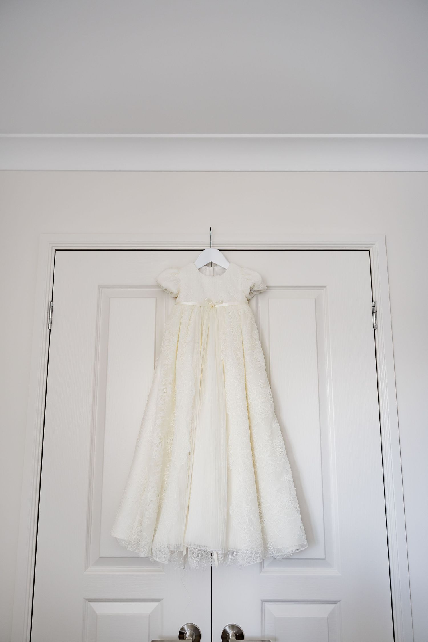 White lace christening gown hanging on a hanger over white double doors.