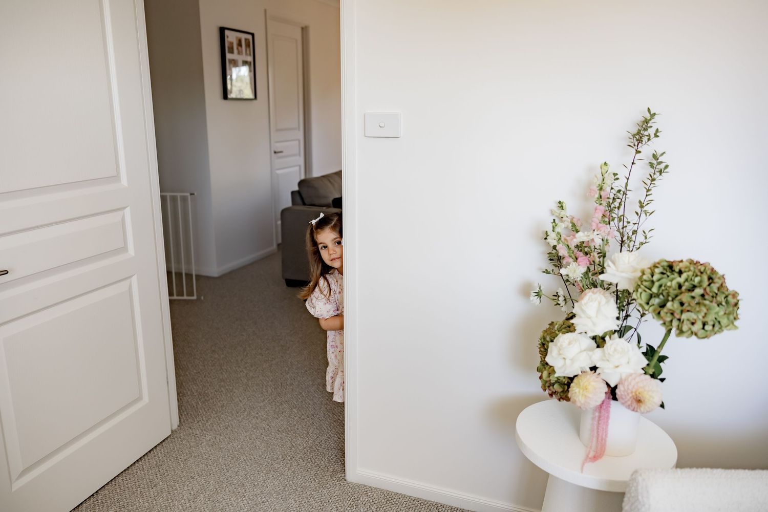 Young girl peeks around white door beside floral arrangement on table in bright hallway.