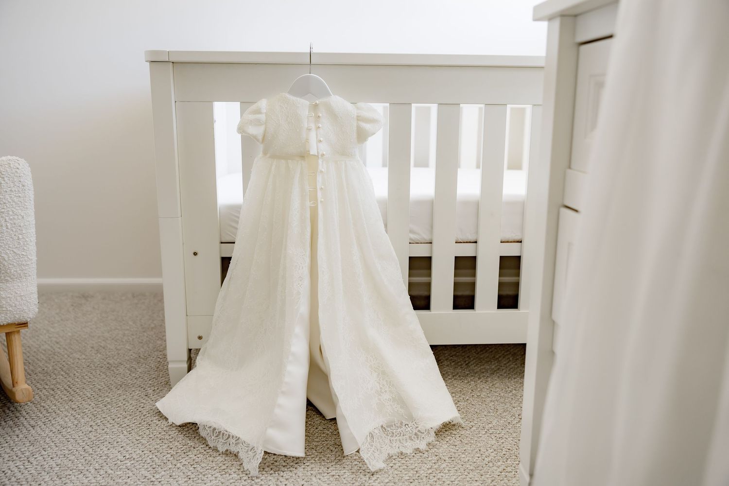 Delicate ivory christening gown with lace trim hanging on hanger in front of white crib.