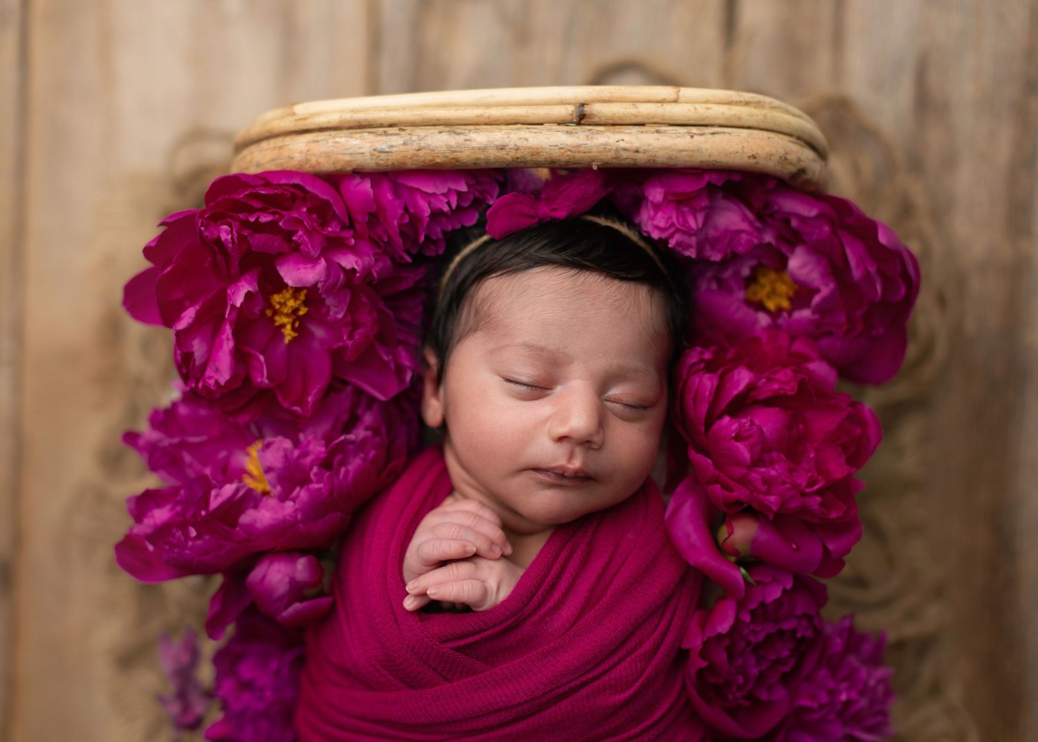 Newborn baby sleeping peacefully in a basket surrounded by vibrant magenta peonies, wrapped in a pink swaddle.