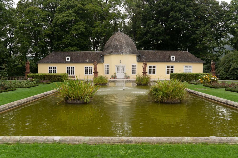 Yellow historic pavilion reflected in a rectangular ornamental pond with manicured hedges.
