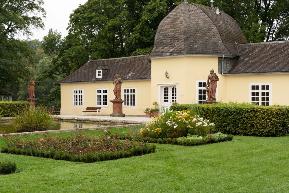 Yellow historic pavilion with statues and manicured garden beds surrounded by lush green trees.