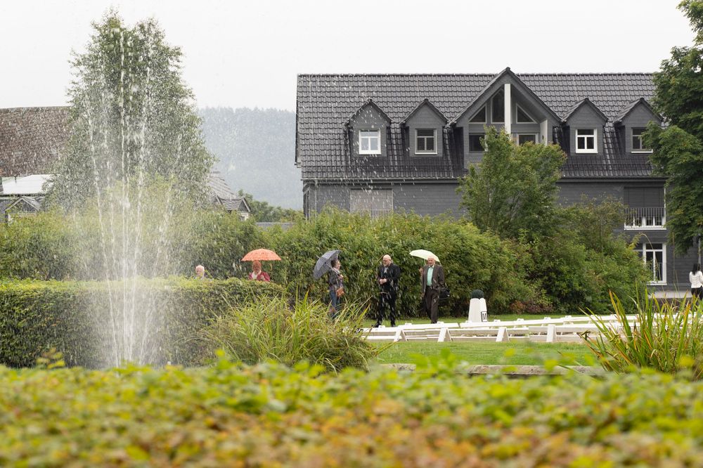 Fountain in a lily pond with people carrying umbrellas walking in the rain near a house.
