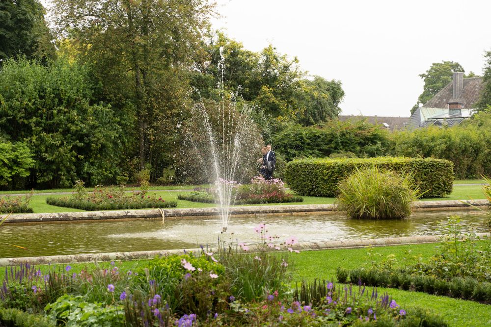 Garden fountain spraying water in an ornamental pond surrounded by colorful flowering plants.