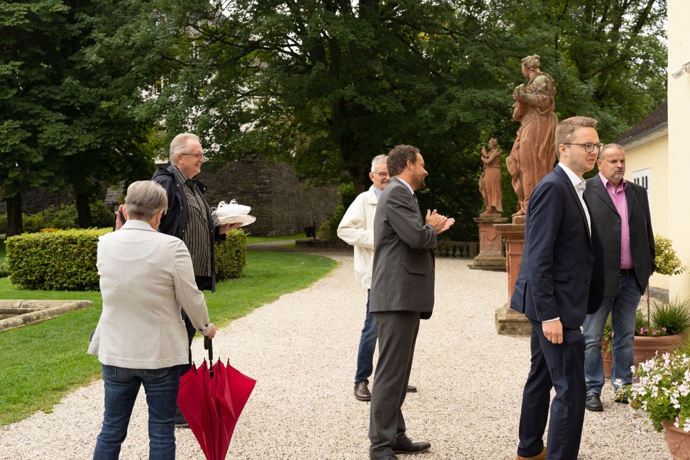 Elegantly dressed guests socializing on a gravel path beside stone statues in a garden.