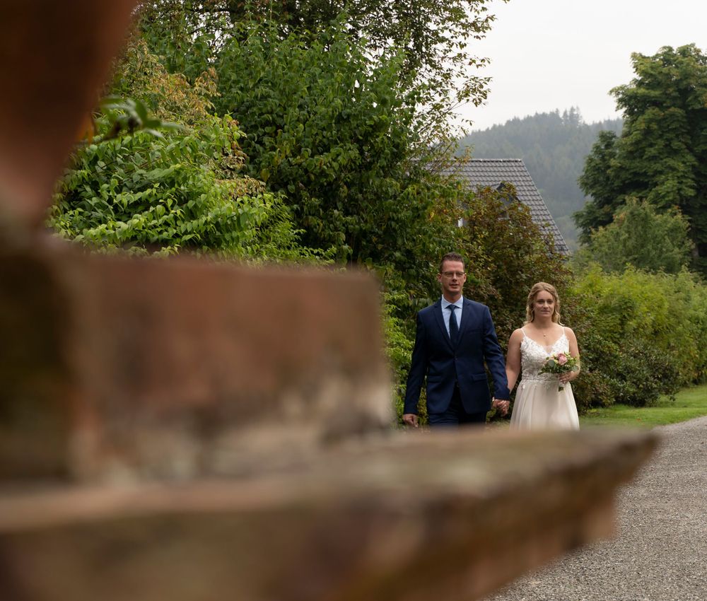 A bride and groom walk hand in hand along a garden path surrounded by lush green trees.