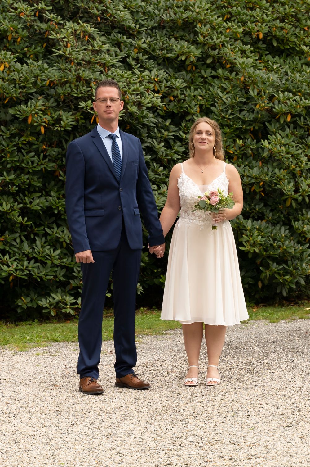 A bride in a white dress holding a bouquet stands with a groom in a navy suit on a gravel path.