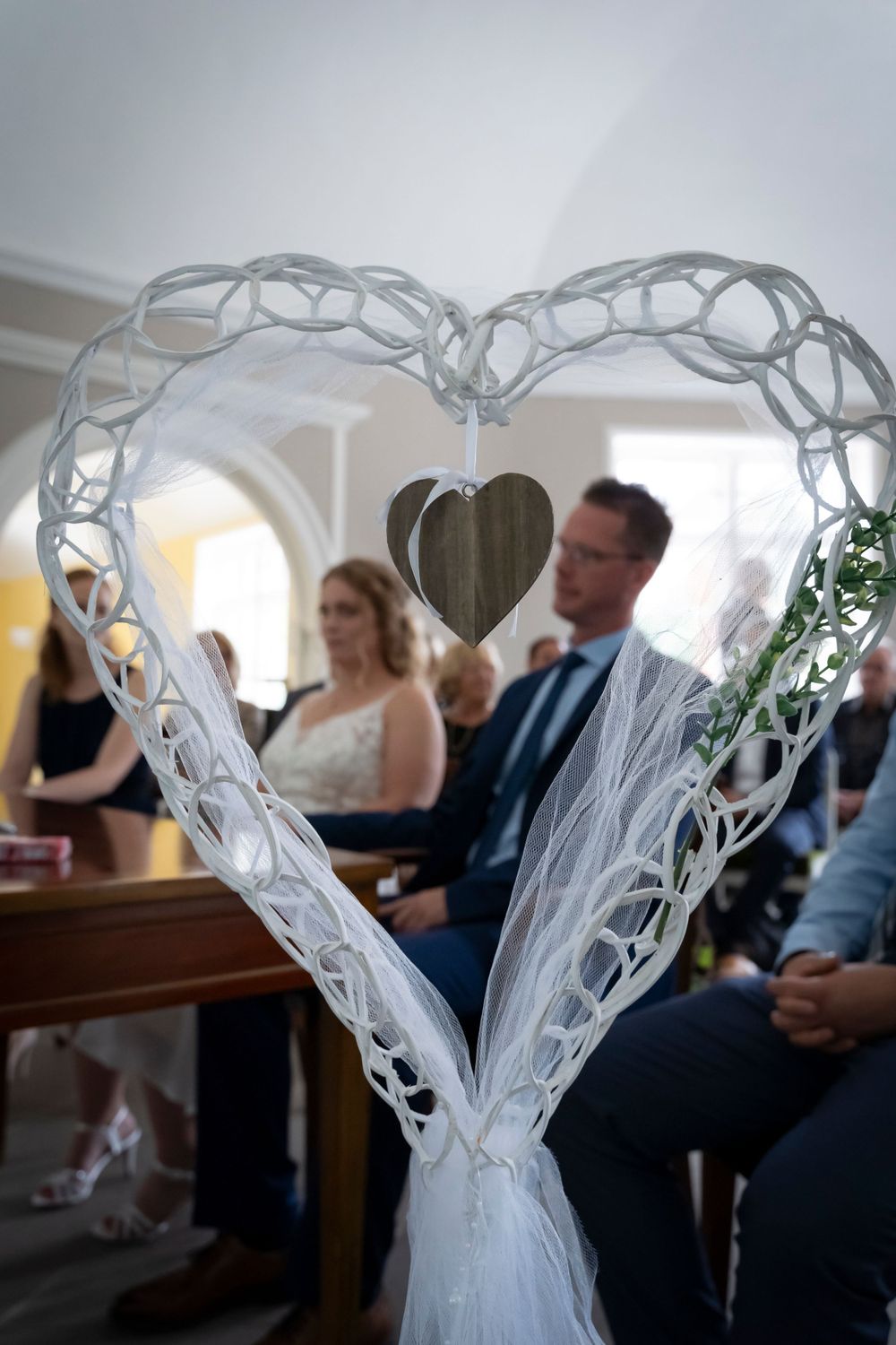 A decorative white heart wreath frames a blurred bride and groom seated at a wedding ceremony.