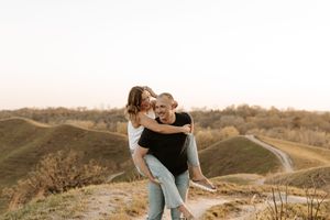 A happy couple enjoys a piggyback ride on a scenic hilltop path at golden hour in a romantic outdoor setting.