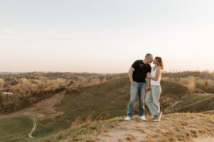 Couple standing close together on a grassy hilltop at golden hour with scenic rolling hills in the background.