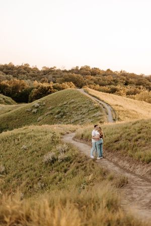Couple embracing on a winding dirt path through rolling green hills at golden hour sunset.