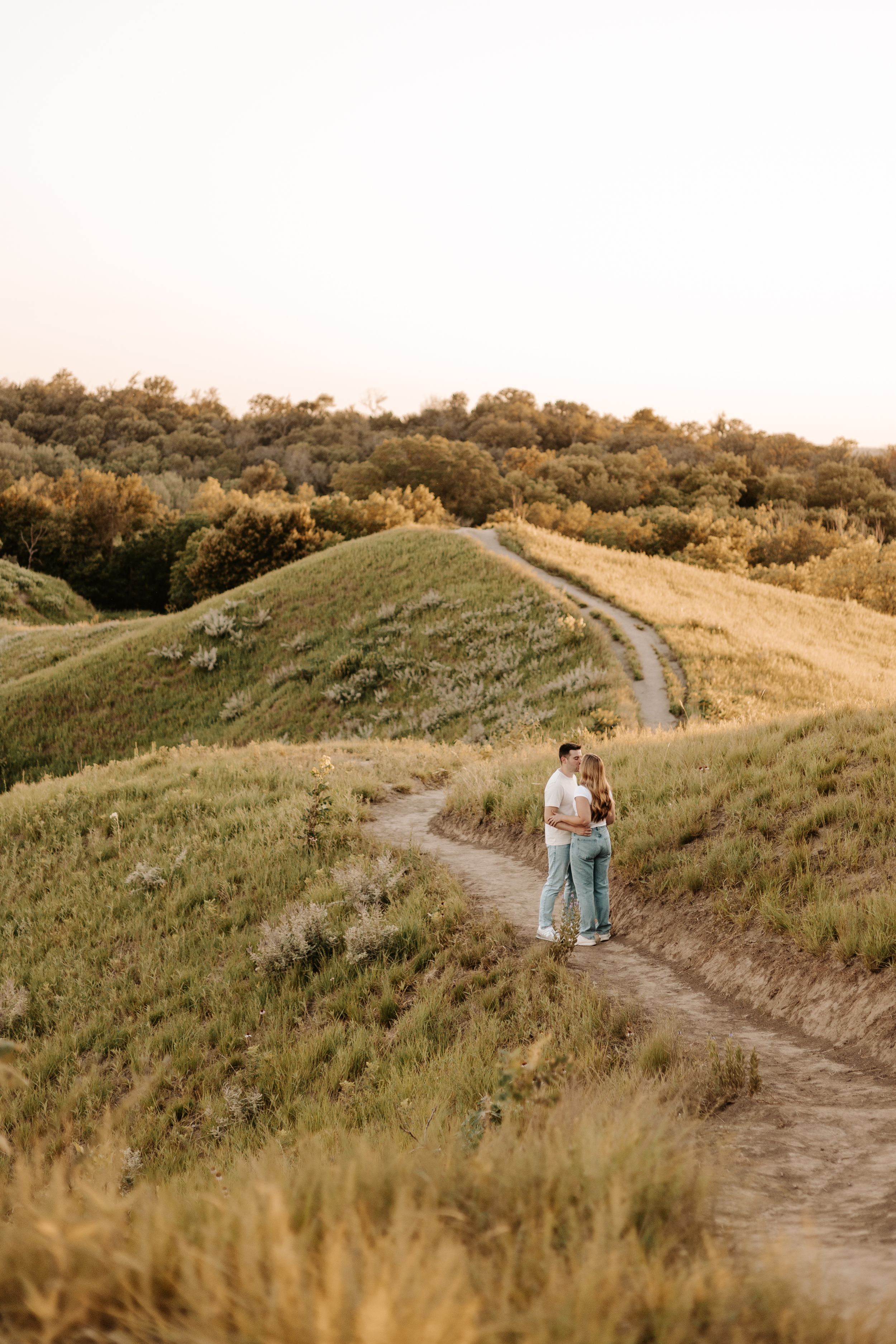 Couple embracing on a winding dirt path through rolling green hills at golden hour sunset.