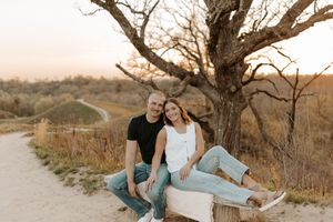Couple sitting on a log at sunset near a dirt path with a bare tree, wearing casual jeans outfits in golden hour light.