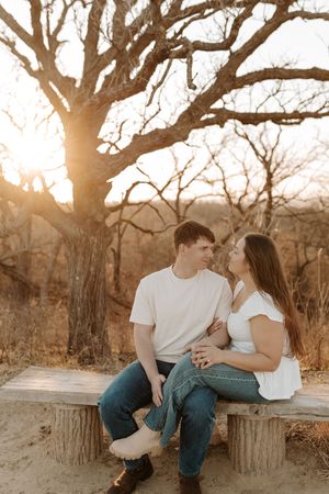 Couple sitting on a rustic bench at golden hour, surrounded by bare trees, sharing a romantic moment outdoors.