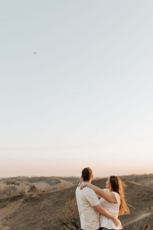 Couple embracing on a hilltop at sunset, gazing at a vast landscape under a soft pastel sky.