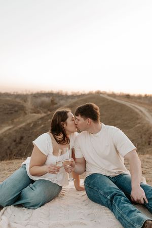 Couple sharing a kiss while holding champagne glasses on a blanket outdoors at sunset.