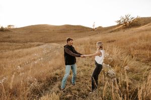 Couple holding hands walking through golden grass fields with rolling hills in the background during autumn.