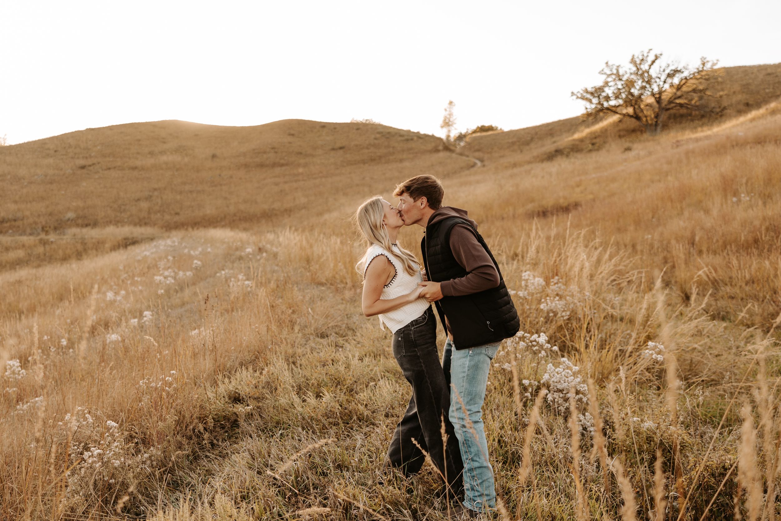 Couple sharing a romantic kiss in a golden meadow with rolling hills and autumn grasses in a scenic outdoor setting.