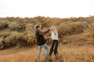 Couple dancing together in golden autumn field with colorful trees in background during sunset.