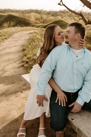 Couple sharing a romantic moment outdoors, woman in white dress kissing man in light blue shirt on a scenic dirt path.