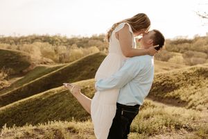 A couple shares a romantic kiss outdoors, man lifting woman in white dress against lush green hills at sunset.