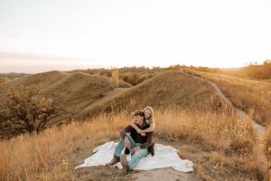 Couple sitting on a white blanket on a golden hillside at sunset, embracing in a romantic outdoor setting.