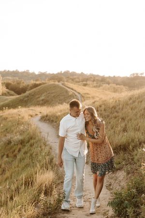 Couple walking hand in hand on a sandy path through golden grass fields during a warm sunset.
