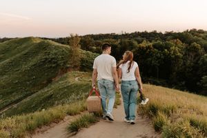 Couple holding hands walking on a scenic trail at sunset, carrying a picnic basket and flowers.