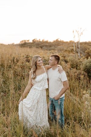 A couple stands in a golden field, the woman in a floral maxi dress smiling at her partner in a white tee and jeans.
